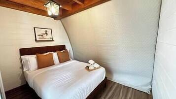 A bedroom with a wooden ceiling, a bed with white bedding, a wooden headboard, a framed picture on the wall, and a bedside table with a lamp.