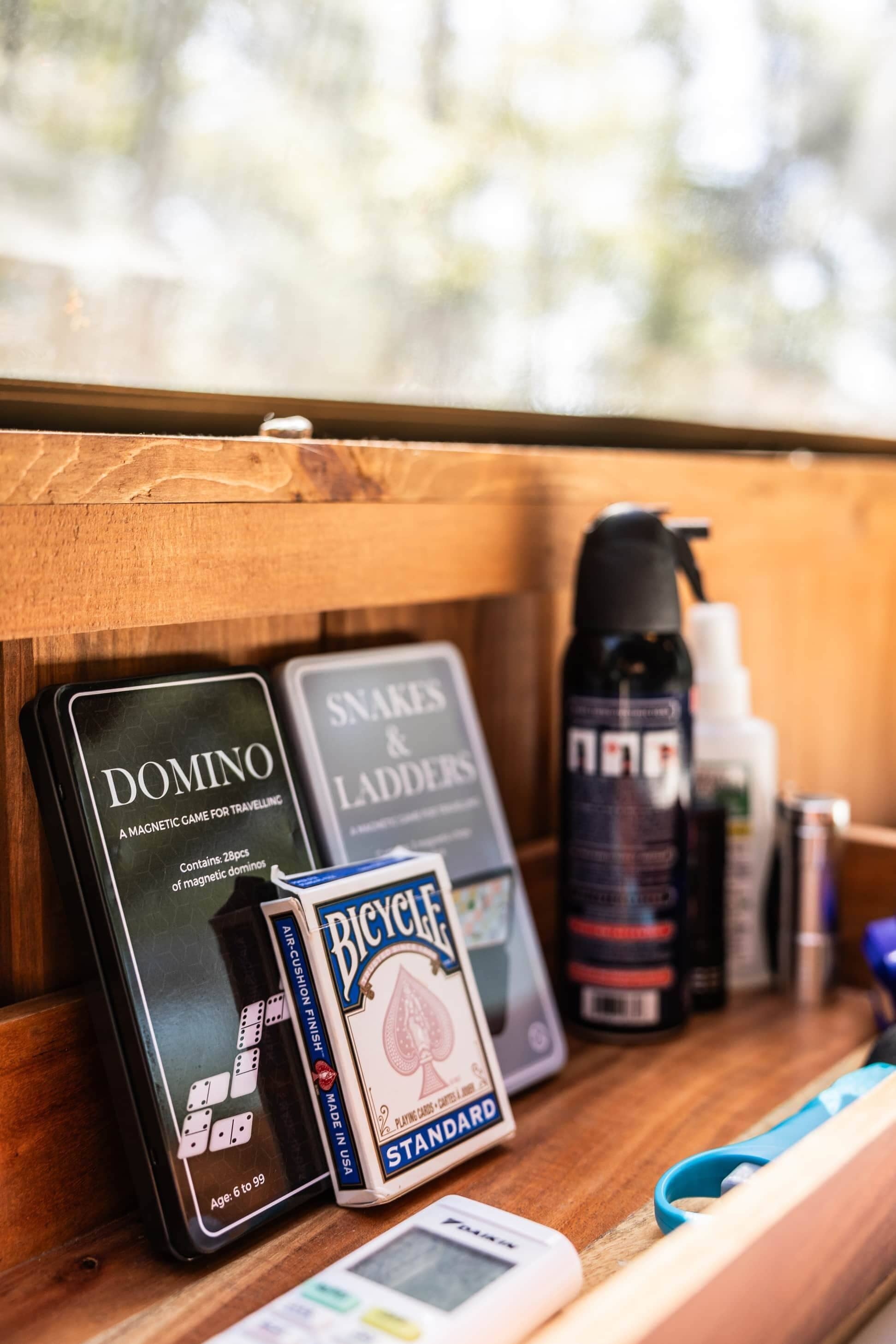 A wooden shelf with a magnetic game called Domino, a game called Snakes & Ladders, a Bicycle playing card deck, and a spray bottle.
