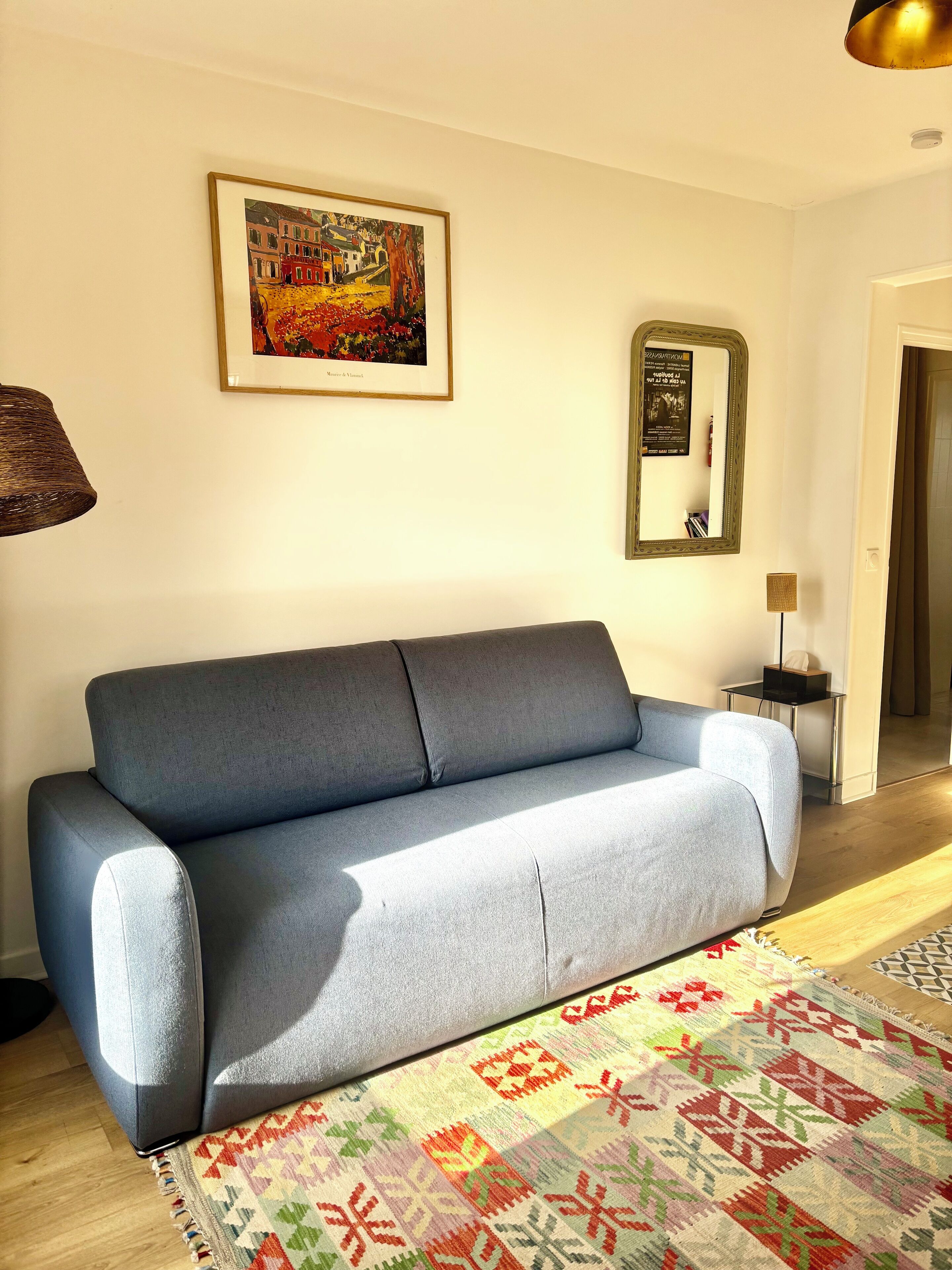 Un salon moderne avec un canapé gris, un tapis coloré, un tableau encadré et un miroir au mur.