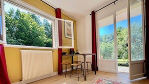 Een kamer met houten vloer, een kamerbreed tapijt met patroon, een tafel met stoelen, een radiator en grote ramen met gordijnen.