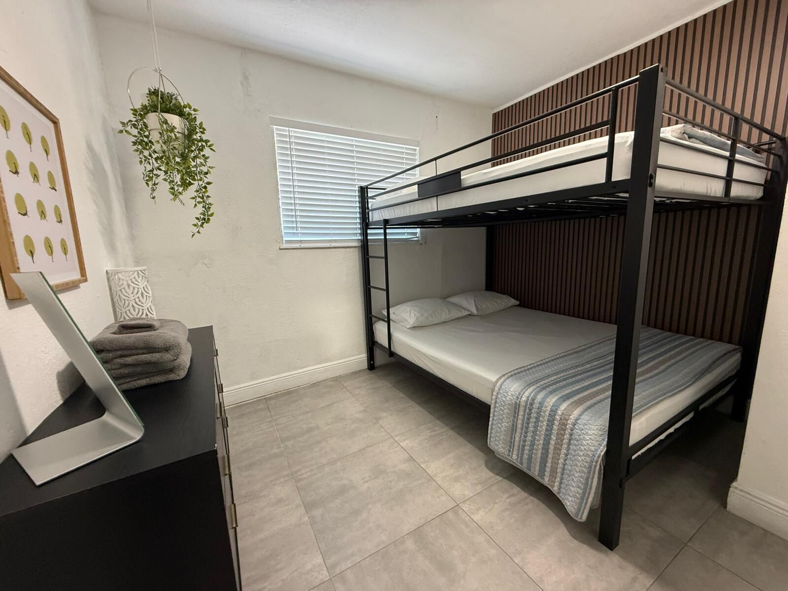 Een slaapkamer met stapelbed, bureau, hangplant en raam met jaloezieën.