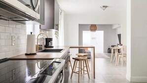 Una cocina moderna con horno empotrado, cafetera y lavamanos. Hay una mesa de comedor de madera con sillas y una lámpara colgante.