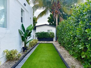 Un sentier étroit et bien entretenu mène à une maison à l’extérieur blanc, entourée d’une végétation luxuriante et d’un palmier.