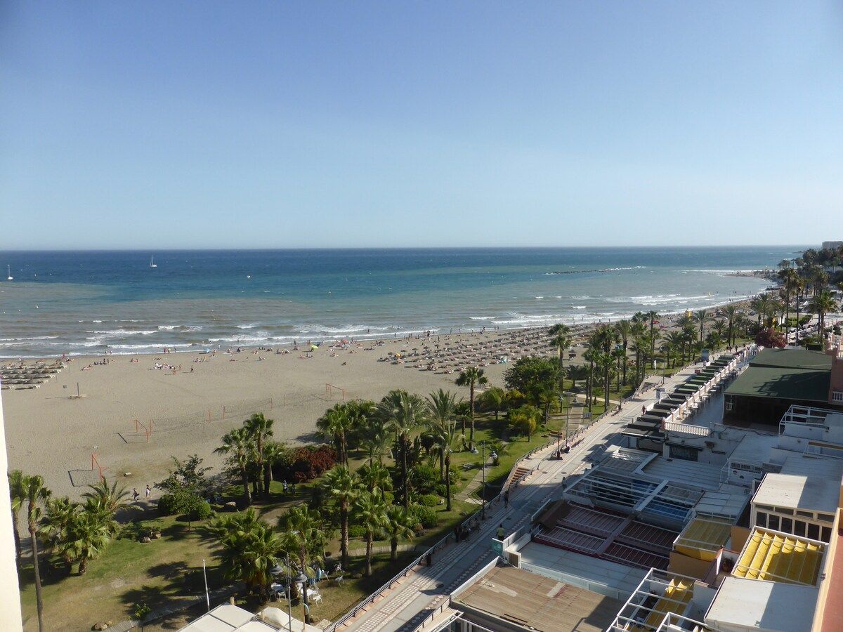 A beach view with a clear sky, a sandy beach, and a row of buildings with a green roof.