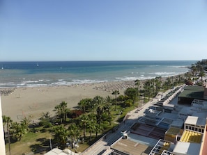 A beach view with a clear sky, a sandy beach, and a row of buildings with a green roof.