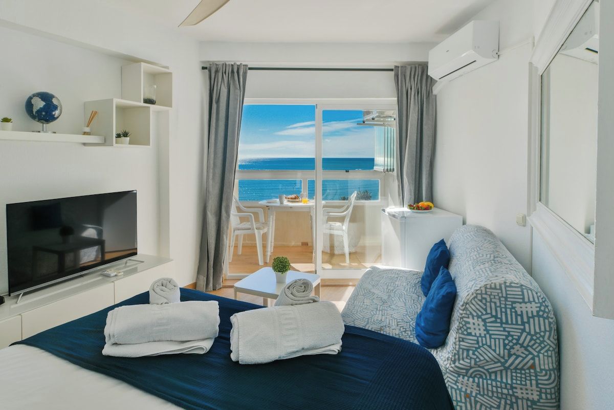 A hotel room with a bed, a TV, a balcony with a view of the sea, and a dining area.