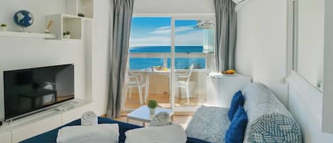 Habitación de hotel con cama, televisión, balcón con vistas al mar y zona de comedor.