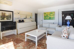 A modern living room with a flat-screen TV, a white coffee table, a grey sofa, and a kitchen area with white cabinets and a painting. - Over the sea - beachfront with amazing views (Fuengirola)