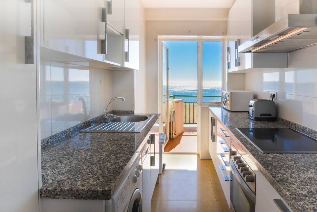 A modern kitchen with granite countertops, stainless steel appliances, and a view of the sea through an open door.