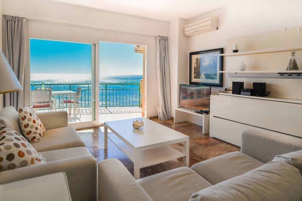 A modern living room with a sofa, coffee table, and TV, a balcony with a view, and a kitchen area.