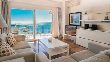 A modern living room with a sofa, coffee table, and TV, a balcony with a view, and a kitchen area.