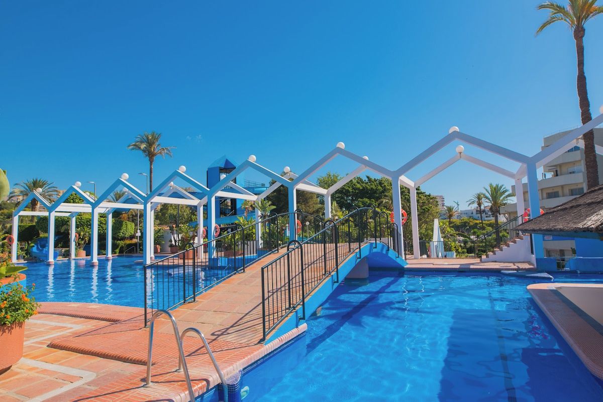 A swimming pool with a geometric canopy structure, surrounded by palm trees and a clear blue sky.