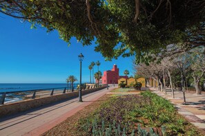 Une promenade en bord de mer, avec un bâtiment rouge, des palmiers et un ciel bleu azur.