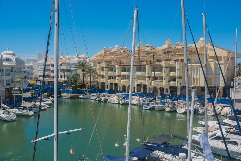 A marina with multiple boats docked, surrounded by buildings with balconies and a clear blue sky.