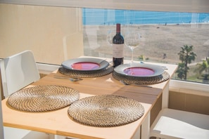 Una mesa con una botella de vino y dos copas, sobre una mesa de madera con individuales tejidos, con vista a una playa.