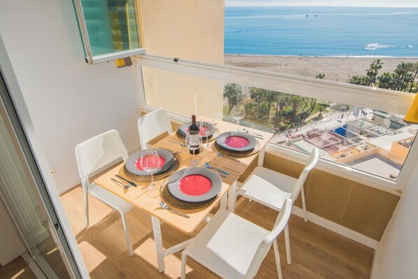 A balcony with a table set for two, overlooking a beach and ocean view.