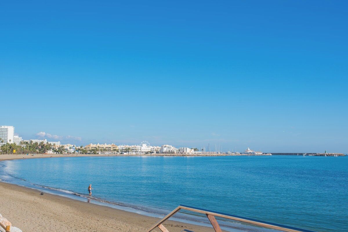 Ein Strand mit blauem Himmel, sandigem Ufer und Gebäuden im Hintergrund.