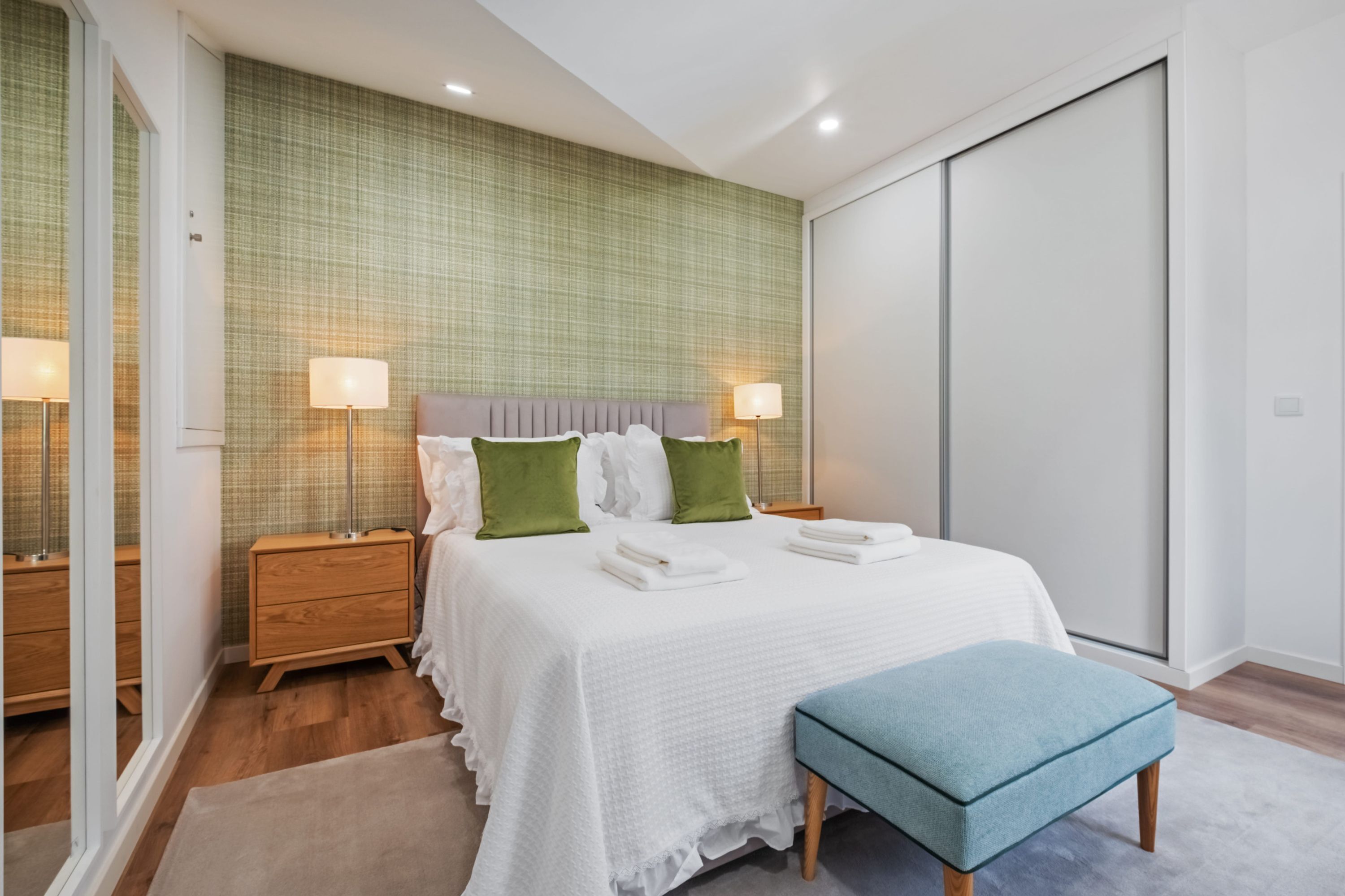A bedroom with a large bed, two bedside tables, a bench, and a sliding door closet.
