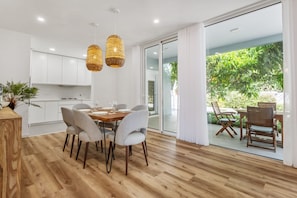 A modern dining area with a wooden table and chairs, a kitchen in the background, and a glass door leading to an outdoor patio.
