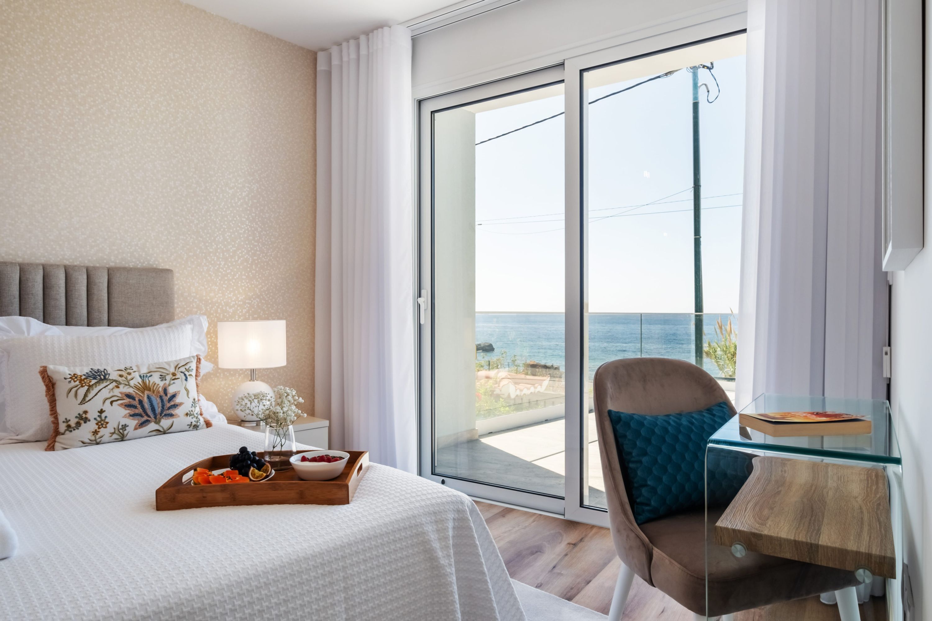 A hotel room with a bed, a nightstand, a glass table, and a view of the ocean.