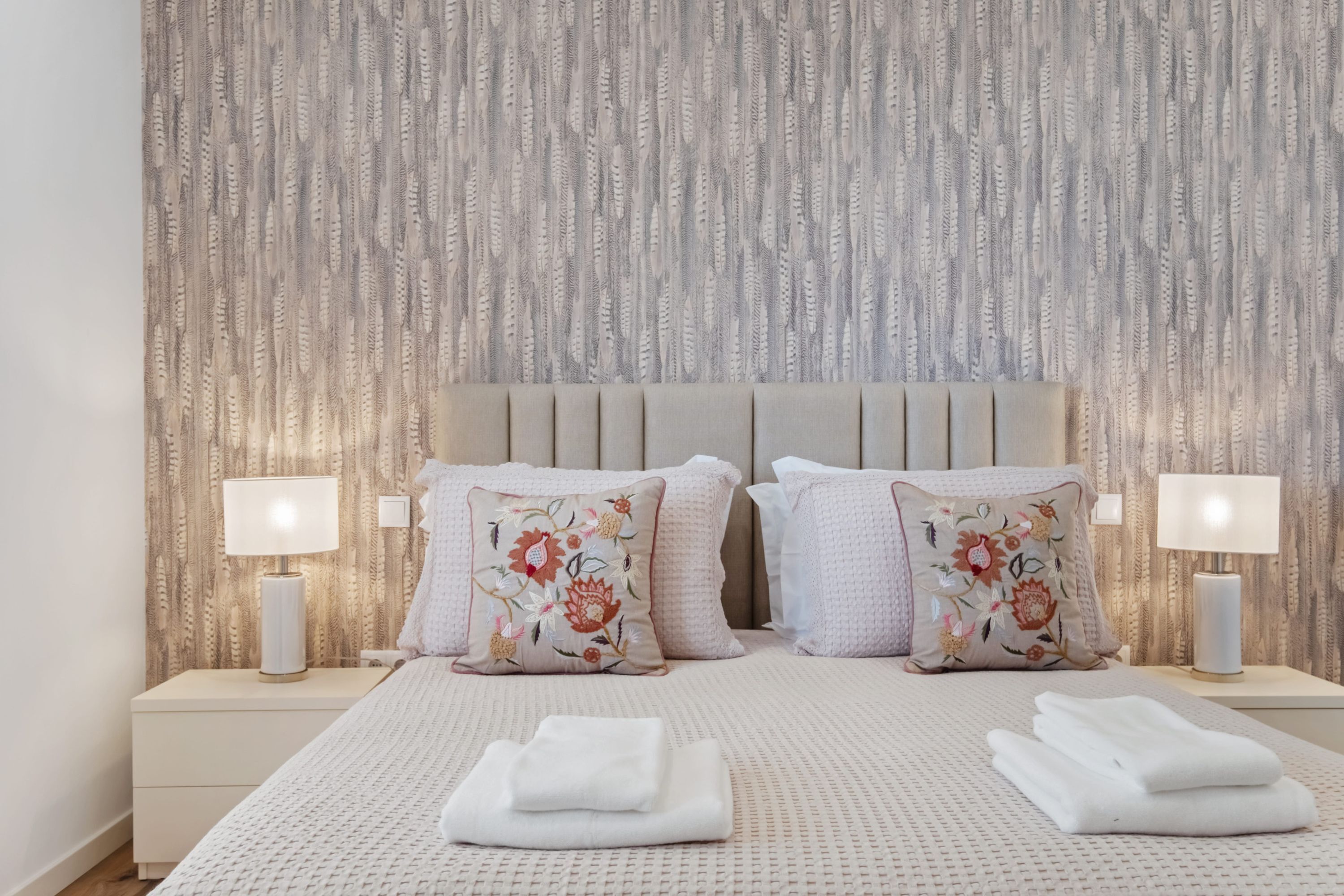 A neatly made bed with a beige headboard, two pillows with floral embroidery, and two bedside lamps on matching nightstands.