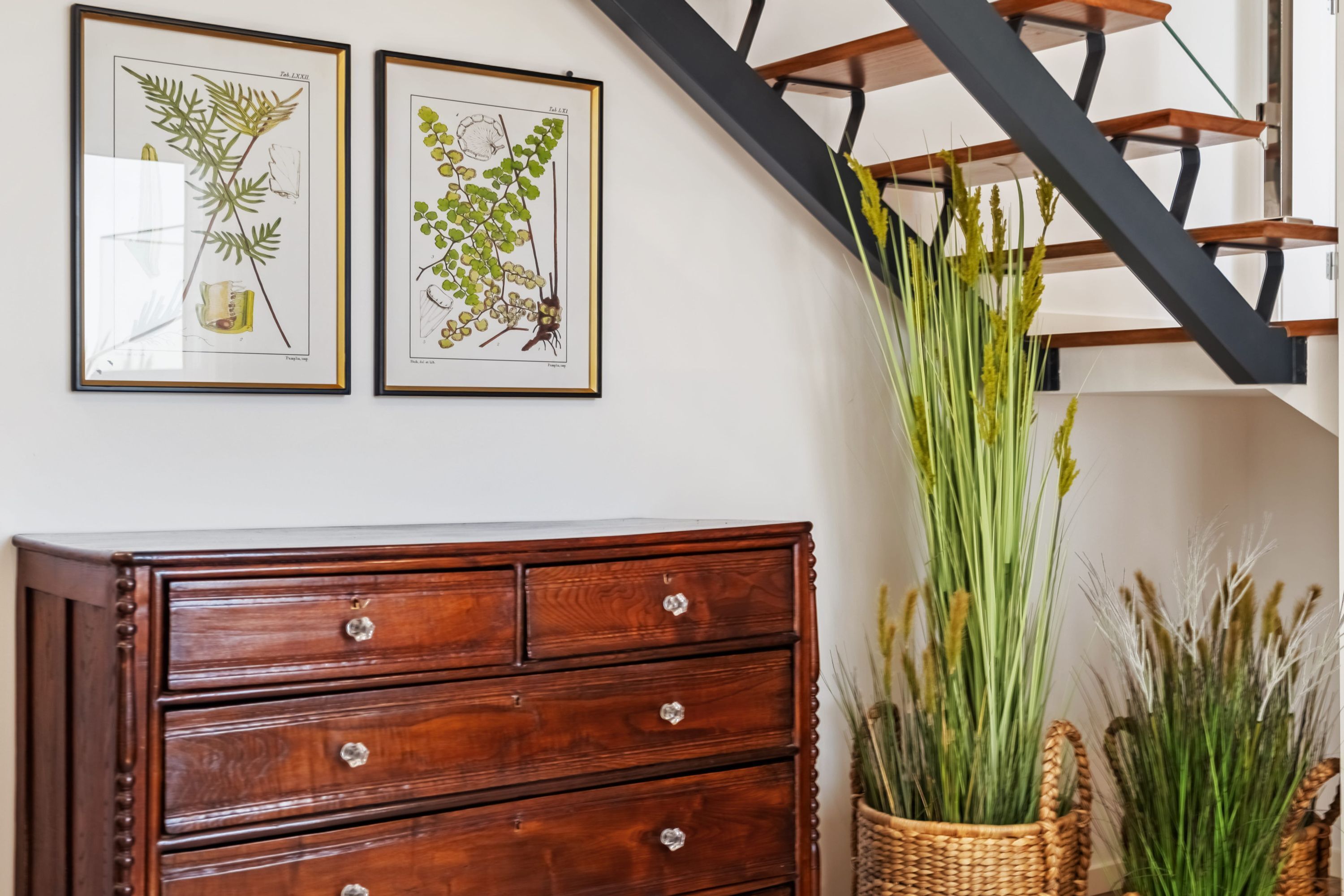 A wooden dresser with drawers, two framed botanical illustrations on the wall, and a staircase with wooden steps.