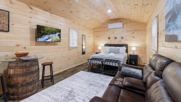 A cozy cabin room with a bed, a leather sofa, a small table with a bowl, and a TV on the wall. - Seneca Hideaway/Sunken Vessel/Hot Tub (Geneva)