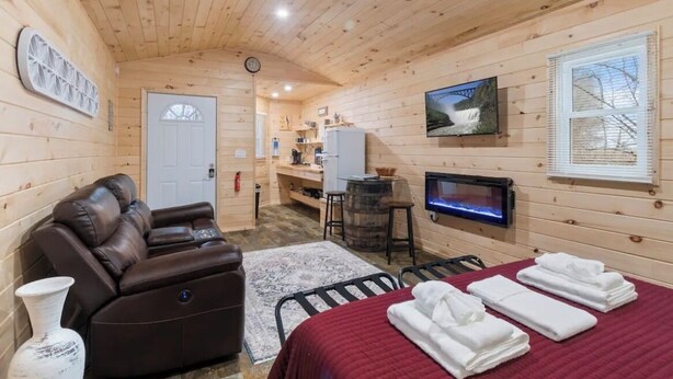 A cozy wooden cabin with a sofa, a bed with towels, a TV, and a fireplace. - Seneca Hideaway/Romantic Getaway Cabin/Hot Tub (Geneva)