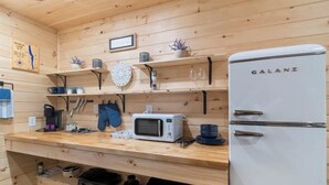 A compact kitchen with wooden walls, a white Galanz refrigerator, a microwave, and shelves holding kitchenware and utensils. - Seneca Hideaway/Romantic Getaway Cabin/Hot Tub (Geneva)