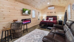 A cozy cabin interior with a wooden ceiling, a television mounted on the wall, a bed with red bedding, a leather sofa, and a rustic barrel. - Seneca Hideaway/Romantic Getaway Cabin/Hot Tub (Geneva)