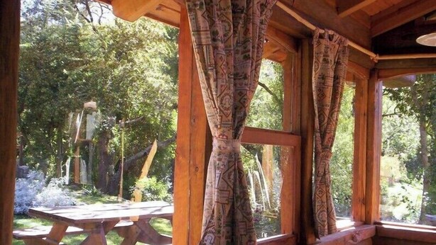 A rustic cabin with large windows and curtains, a wooden ceiling, and a view of a picnic table and greenery outside. - Apart Suites & Cabañas Duendes del Pungo (La Cumbre)