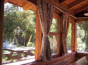 Un chalet rustique doté de grandes fenêtres et de rideaux, d’un plafond en bois et offrant une vue sur une table de pique-nique et la verdure environnante.
