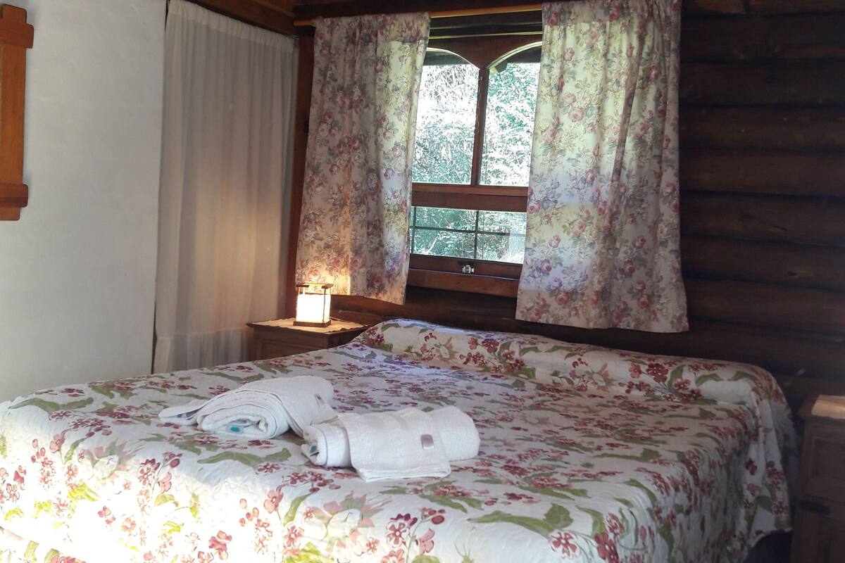 Une chambre dans un chalet en bois, avec un lit, une table de chevet dotée d’une lampe et une fenêtre ornée de rideaux à motifs floraux.