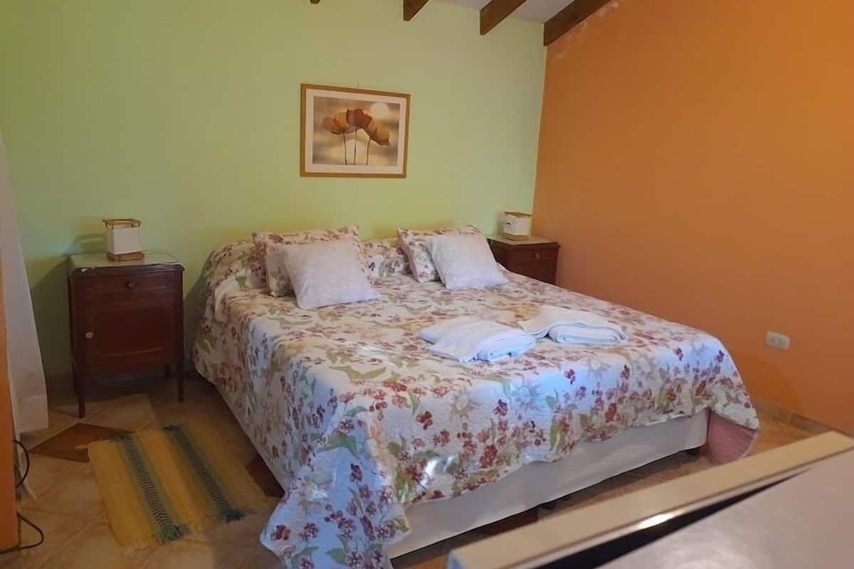 Une chambre à coucher avec une couvre-lit à motifs floraux, deux tables de chevet, une petite table d’appoint, un tableau encadré au mur et un tapis rayé sur le sol.