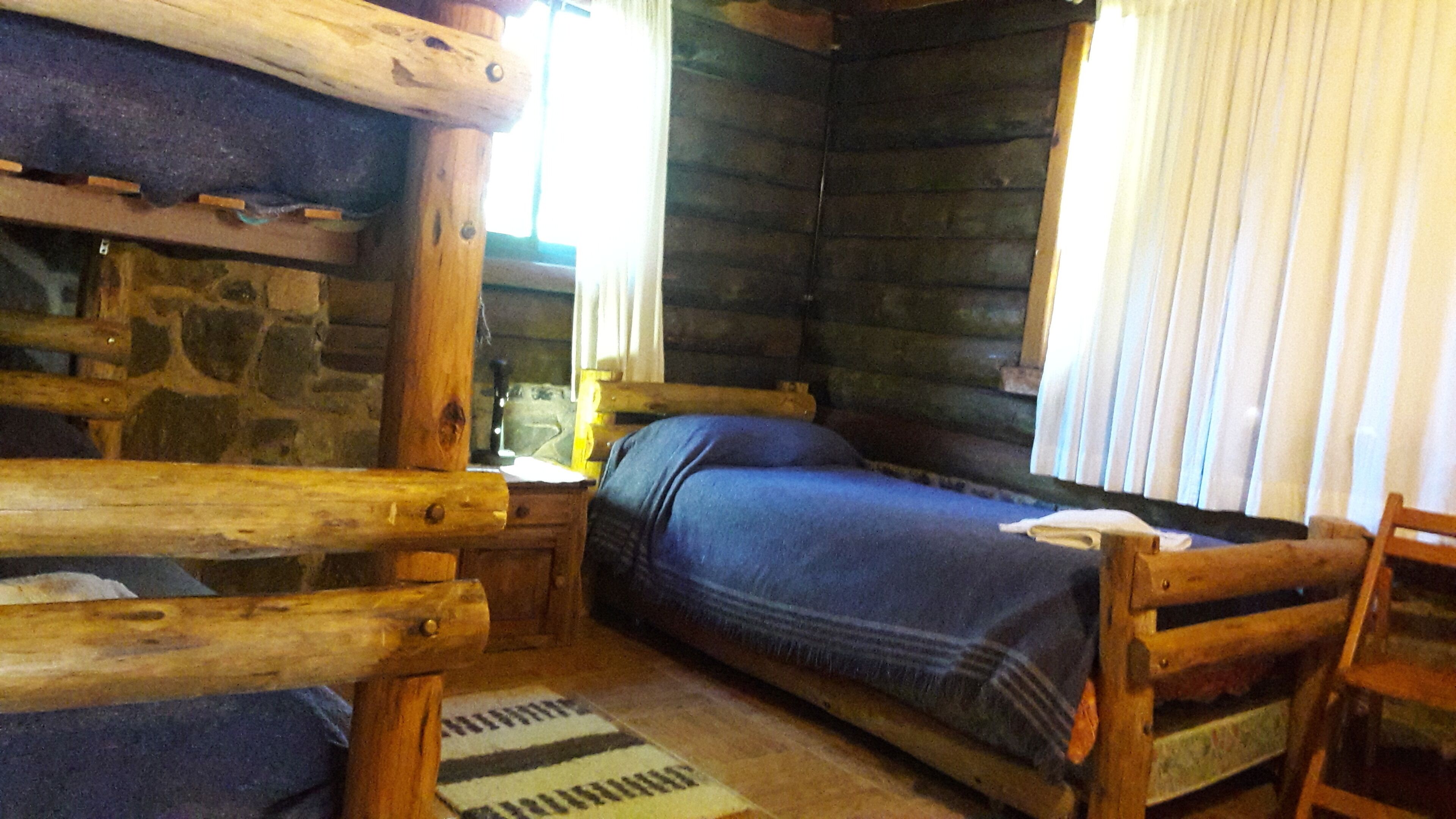 Quarto de cabana de madeira com cama, paredes de madeira, lareira de pedra e janela com cortinas.