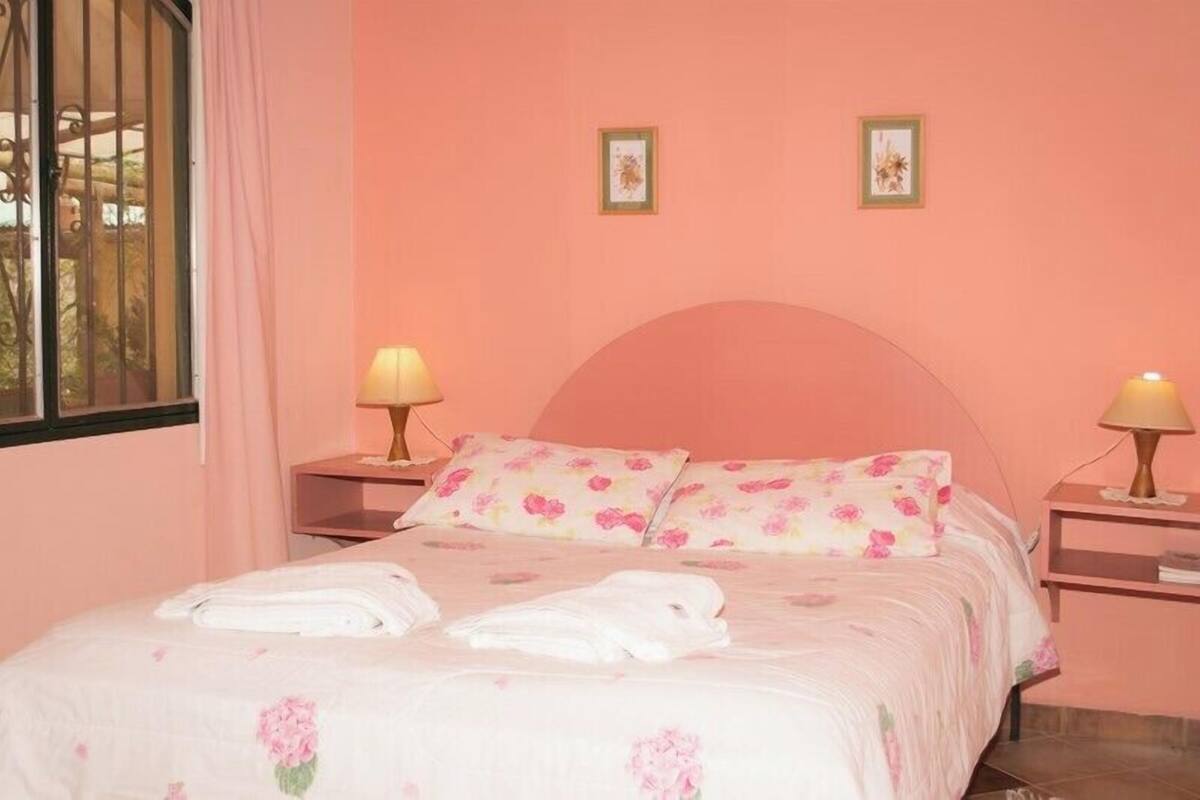 Une chambre avec un mur rose, une literie à motifs floraux, deux lampes de chevet et une fenêtre avec un rideau.