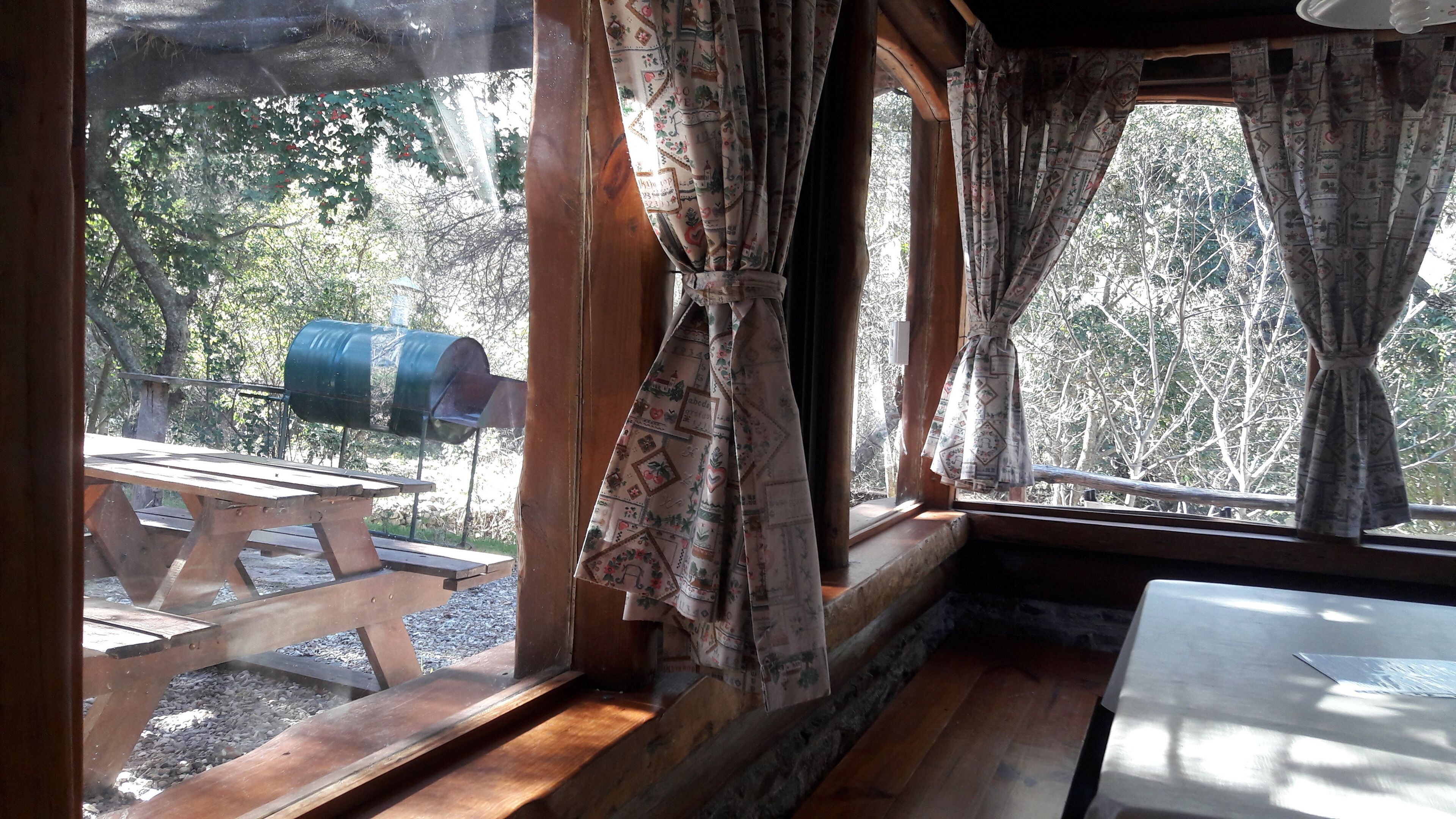 Interior rústico de una cabaña con un banco de madera afuera, una mesa adentro y vistas a árboles y un barril a través de la ventana.