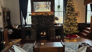 A cozy living room with a stone fireplace, a Christmas tree, and a television displaying a snowy scene.