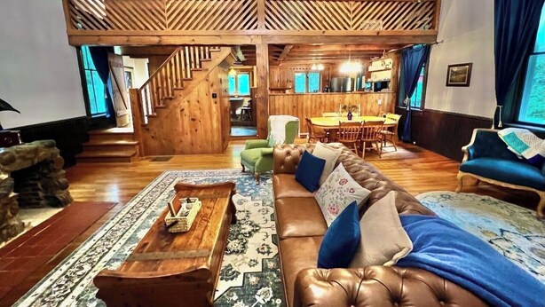 A spacious living area with a high wooden ceiling, a staircase leading to an upper level, a dining area, and a cozy seating arrangement.