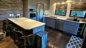 A modern kitchen with a large island, dark wooden flooring, stainless steel appliances, and a stone wall backsplash. - Lake Loft at Deep Creek Lake (Oakland)