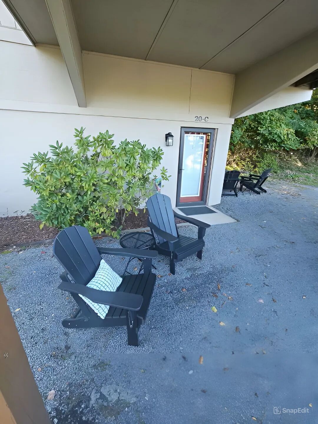 Un espace extérieur aménagé avec des chaises Adirondack et une table, une porte portant le numéro 20-C, et un petit arbuste en arrière-plan.