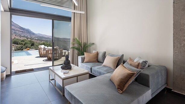 A modern living room with a grey sofa, a small table, and a view of a pool and mountains. - Agrimi Luxury Suite with Private Pool  (Sfakia)