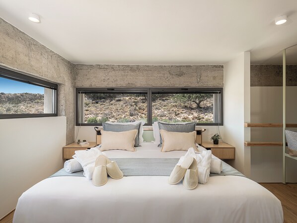 A bedroom with a large bed, two nightstands, a window with a view of a desert landscape, and a wooden floor.