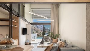 A modern living room with a grey sofa, a coffee table, and a view of a mountainous landscape through large windows. - Agrimi Luxury Suite with Private Pool  (Sfakia)
