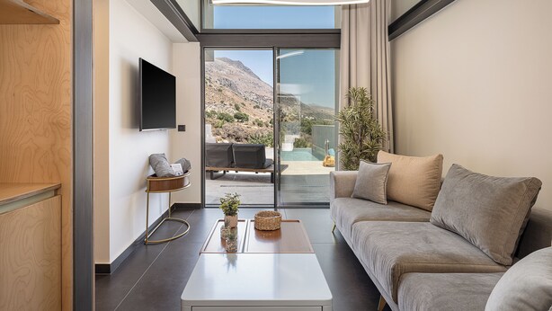 A modern living room with a sofa, coffee table, and a TV. There is a view of a mountainous landscape through large glass doors.