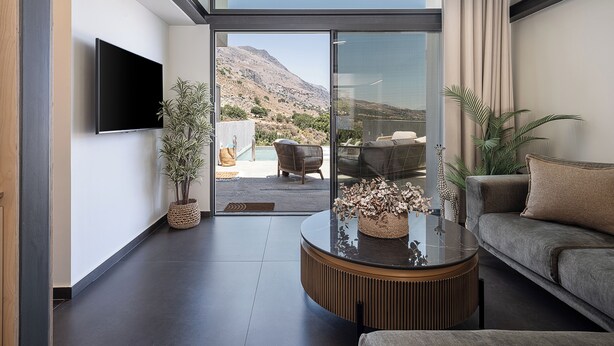 A modern living room with a flat-screen TV, a round coffee table, and a sofa. There's a large window offering a view of a mountainous landscape.