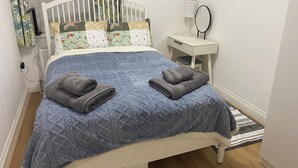 A neatly made bed with a blue quilt and two folded towels, a white bedside table with a mirror, and a small desk with a lamp. - Slough - Sleeps 3 - Petfriendly - Riverside - Bay (Slough)