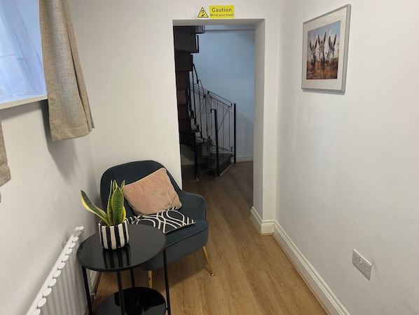 A hallway with a staircase, a black chair with a cushion, a small table with a plant, and a framed picture on the wall. - Slough - Sleeps 3 - Petfriendly - Riverside - Bay (Slough)
