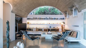 A modern living room with a white sofa, two black chairs, a coffee table, and a stone sculpture. A large arched window provides a view of greenery outside. - Villa Luna by MyVillaCollection (Maiori)