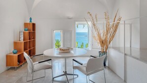 A modern room with a round table, white chairs, a wooden bookshelf, and a view of the sea through a window. - Villa Luna by MyVillaCollection (Maiori)